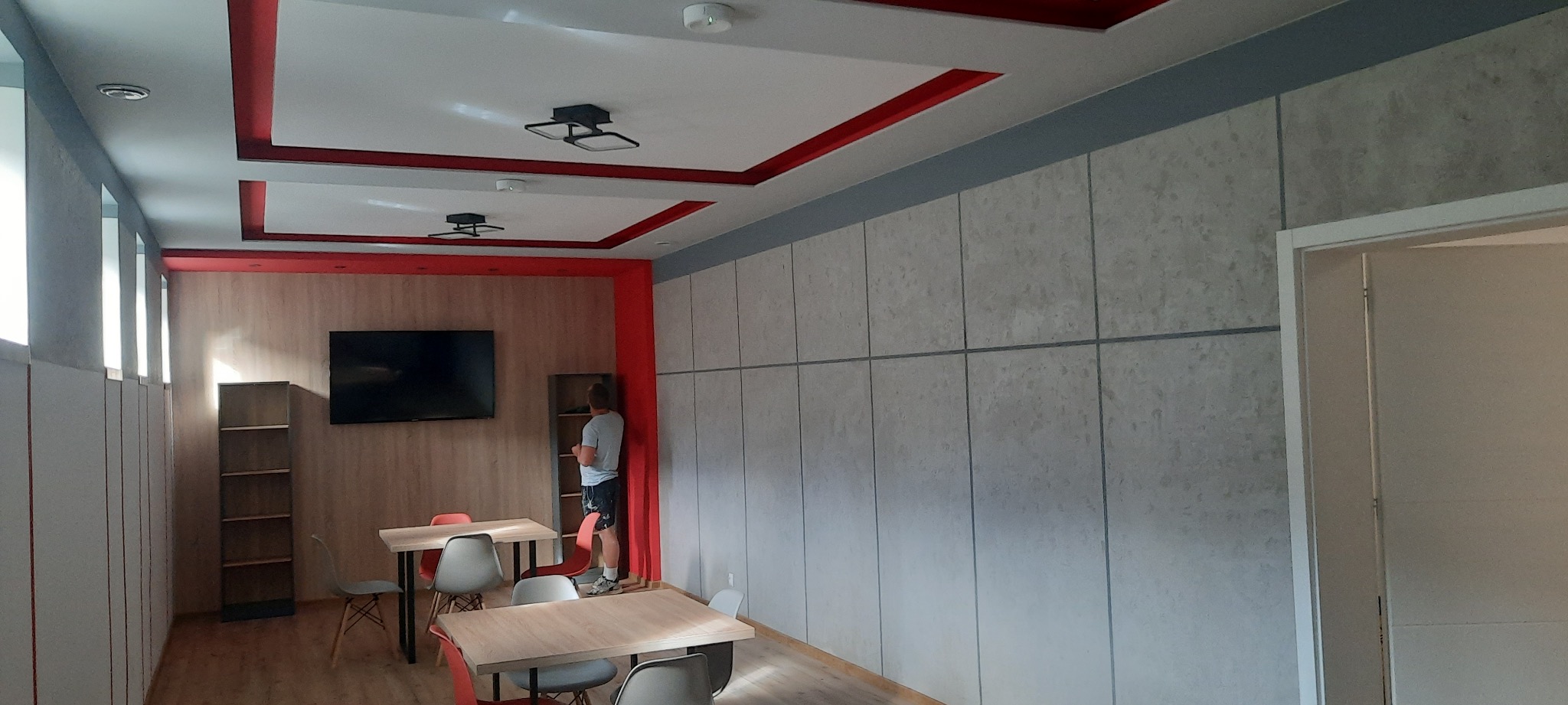 Nowoczesne wnętrze biura z panelami ściennymi w odcieniach szarości, czerwonymi akcentami na suficie i drewnianymi meblami, oświetlone nowoczesnymi lampami sufitowymi.