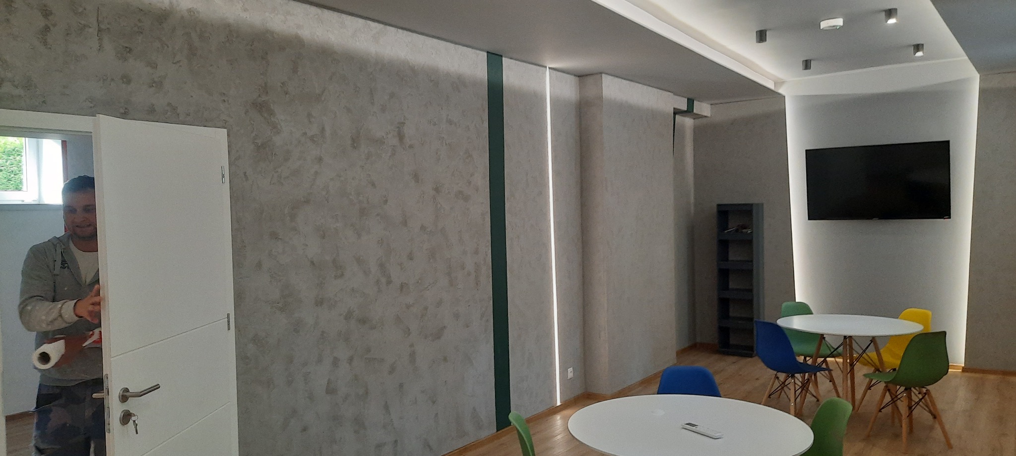 Mężczyzna z rolką taśmy malarskiej w ręku, wchodzący przez białe drzwi do nowocześnie wykończonego biura z dekoracyjną fakturą na ścianach, podświetleniem LED i kolorowymi krzesłami wokół białych...