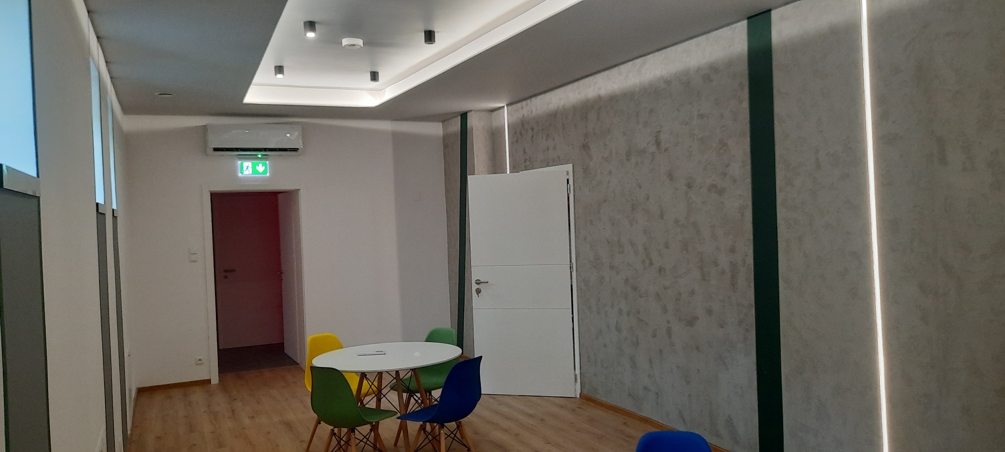 Wykończone wnętrze biura z dekoracyjną ścianą o fakturze betonu, podświetloną pionowymi paskami LED, białe drzwi i okrągły stół z kolorowymi krzesłami.