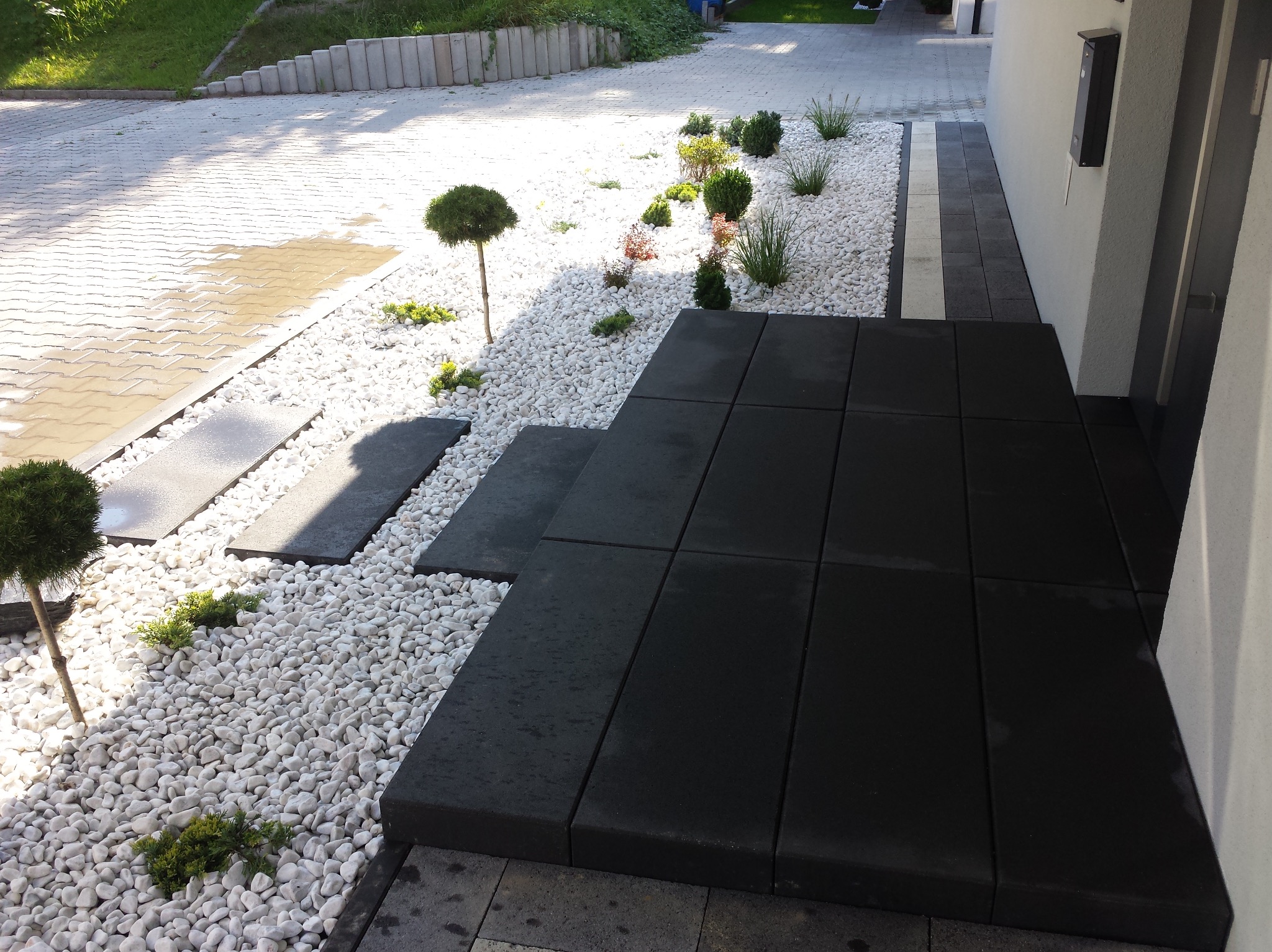 Nowoczesne wejście do domu z czarnych płyt tarasowych, otoczone białymi kamieniami i ozdobnymi roślinami, prowadzące do drzwi wejściowych.