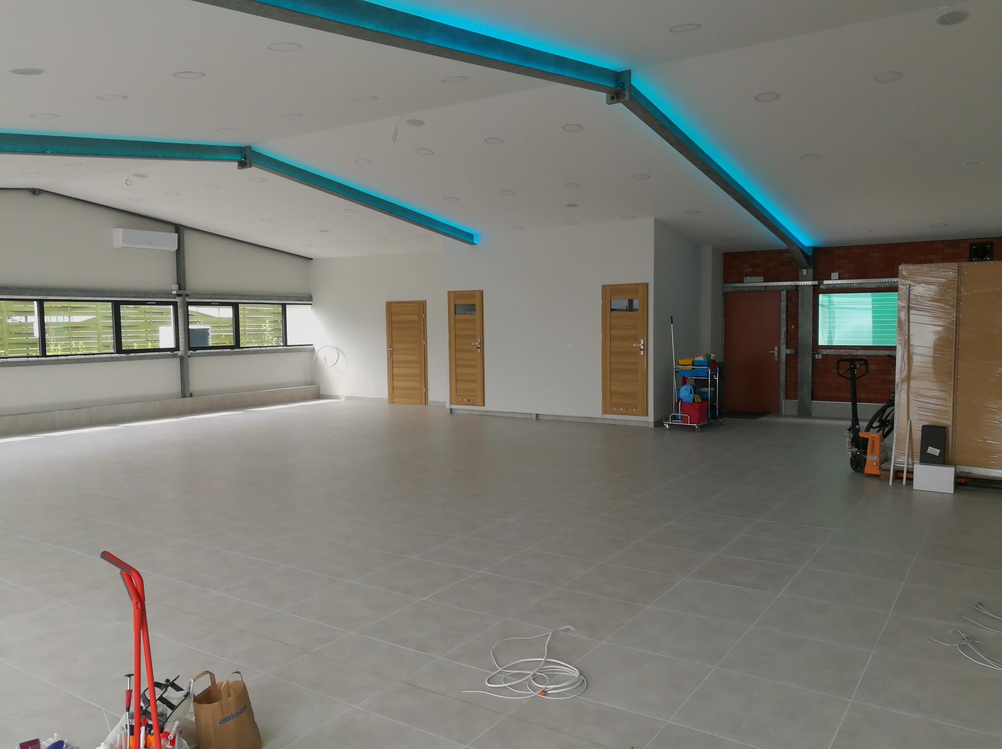 Przestronne, jasne wnętrze hali w Nowym Sączu z szarą, wykafelkowaną podłogą, trzema drewnianymi drzwiami, oświetleniem LED w suficie, oknami po lewej stronie i sprzętem budowlanym w tle.