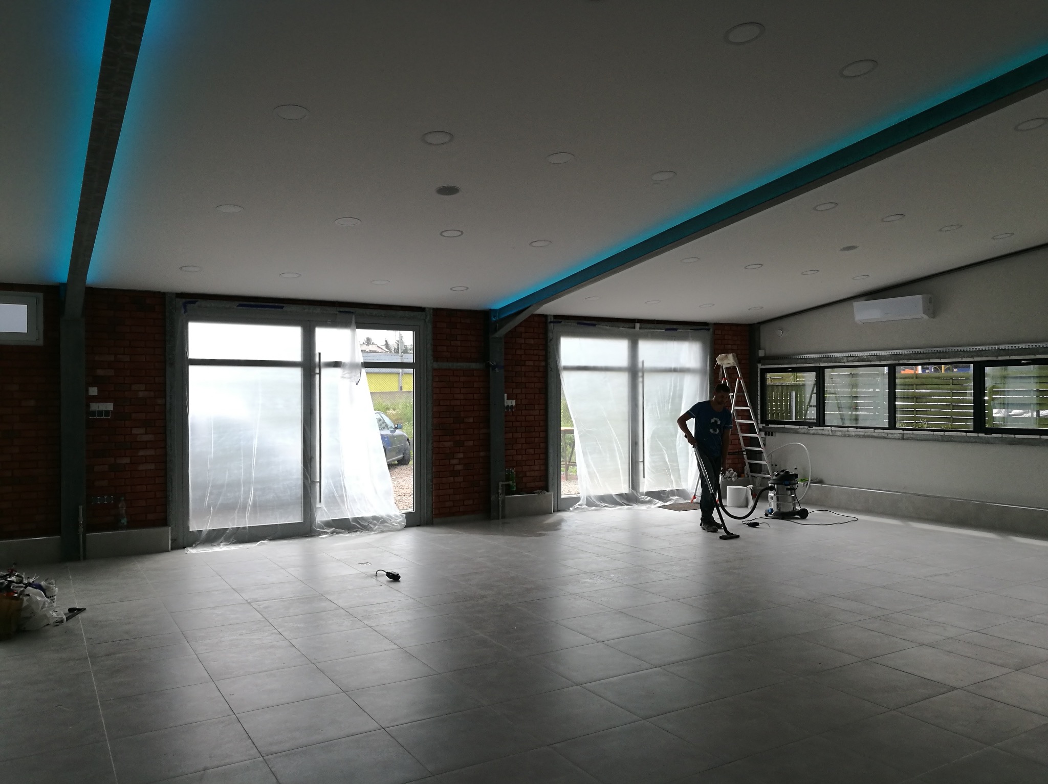 Przestronne wnętrze z szarą podłogą z płytek, osłoniętymi folią oknami i pracownikiem sprzątającym odkurzaczem przemysłowym, widoczne elementy ceglanej ściany i oświetlenie LED na suficie.