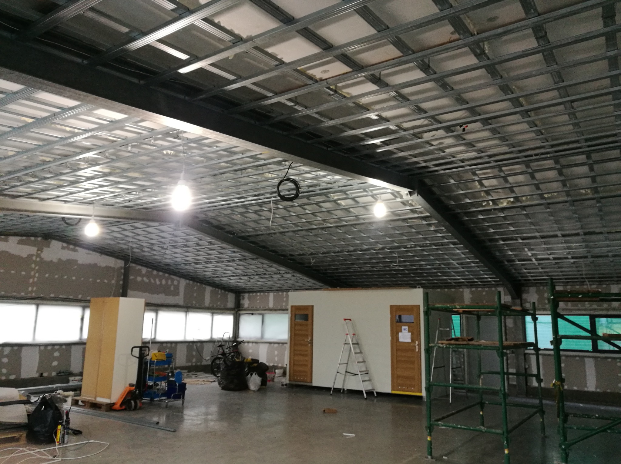 Wnętrze hali w trakcie prac wykończeniowych: widoczna metalowa konstrukcja sufitu podwieszanego, ściany w trakcie szpachlowania, przenośna kabina sanitarna z drewnianymi drzwiami, rusztowania...