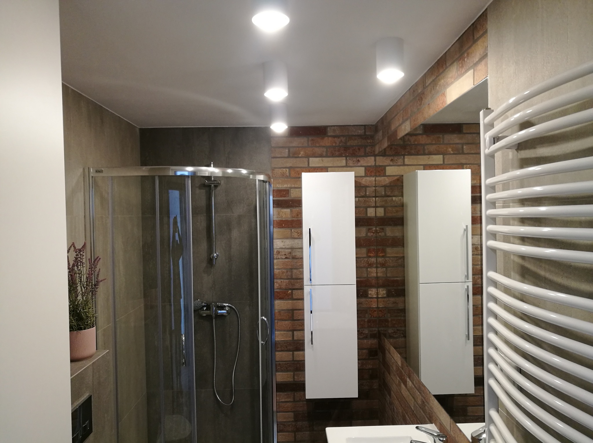 Nowoczesna łazienka z kabiną prysznicową, białymi szafkami wiszącymi na ścianie z cegły i grzejnikiem drabinkowym.