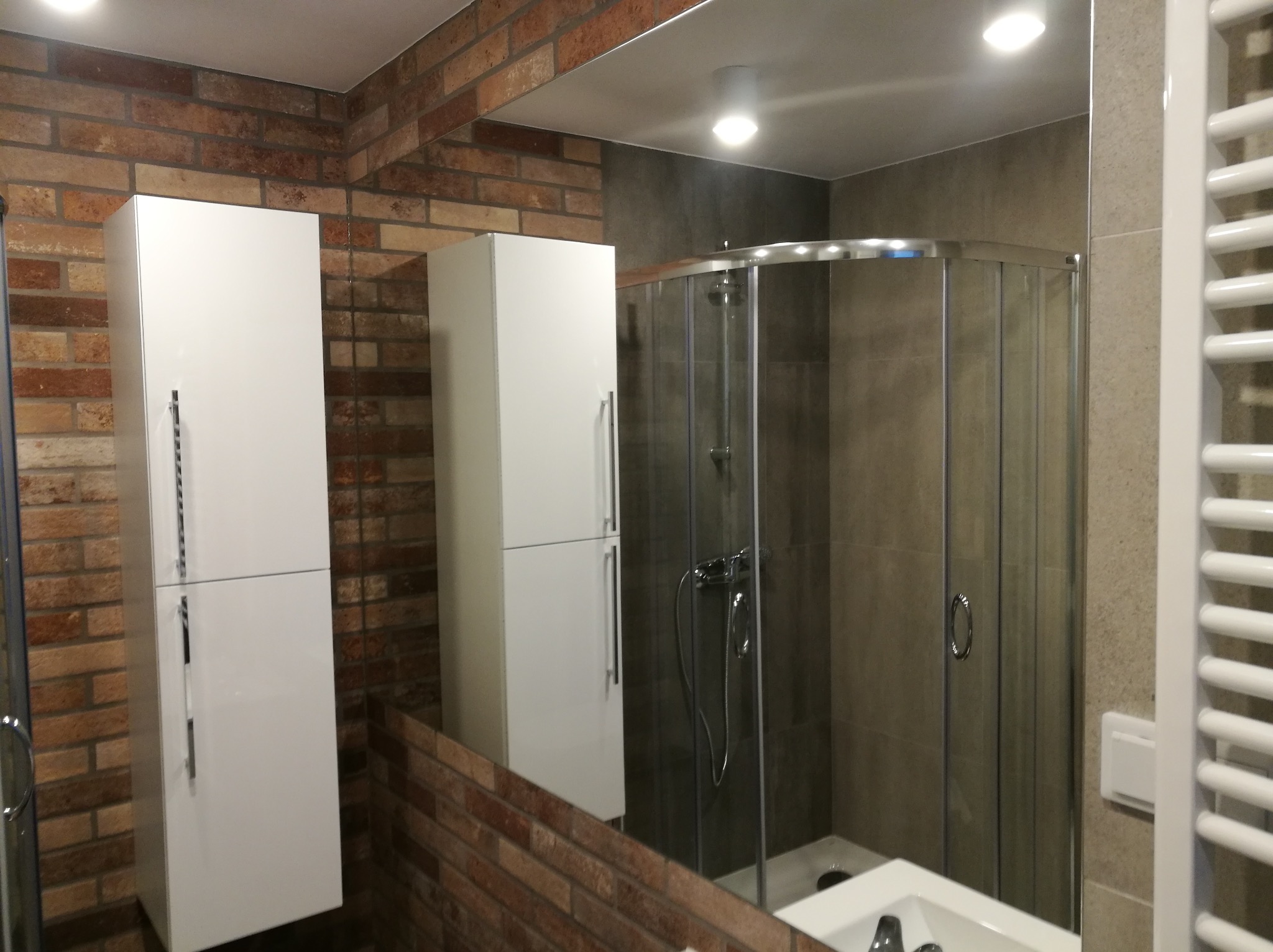 Nowoczesna łazienka z prysznicem w kabinie z przesuwanymi drzwiami, umywalką wpuszczaną w blat, białymi szafkami wiszącymi z długimi uchwytami i ścianą wyłożoną cegłą.