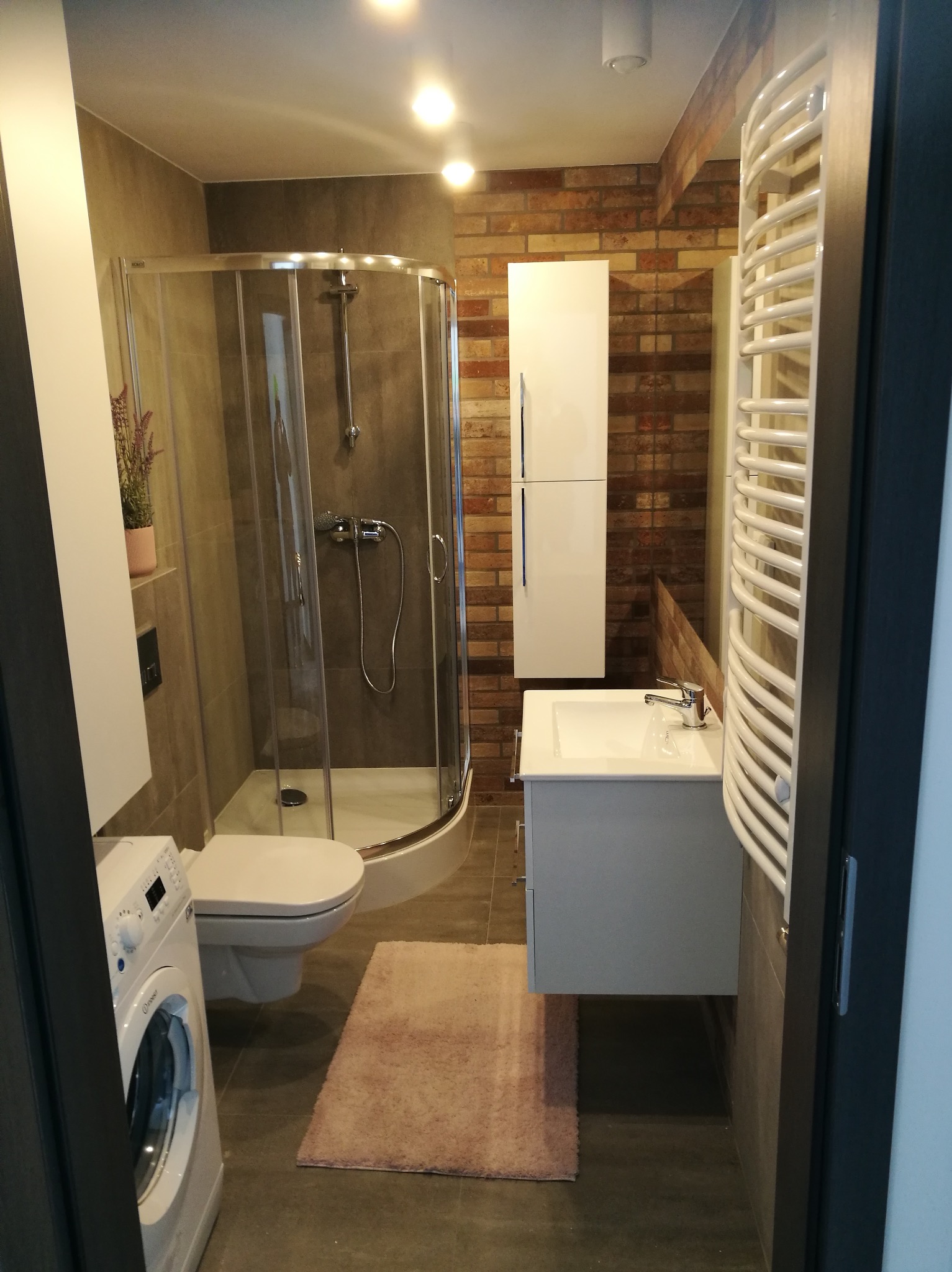 Nowoczesna łazienka z kabiną prysznicową, wiszącą szafką z umywalką, grzejnikiem drabinkowym, pralką i dekoracyjną ścianą z cegły.