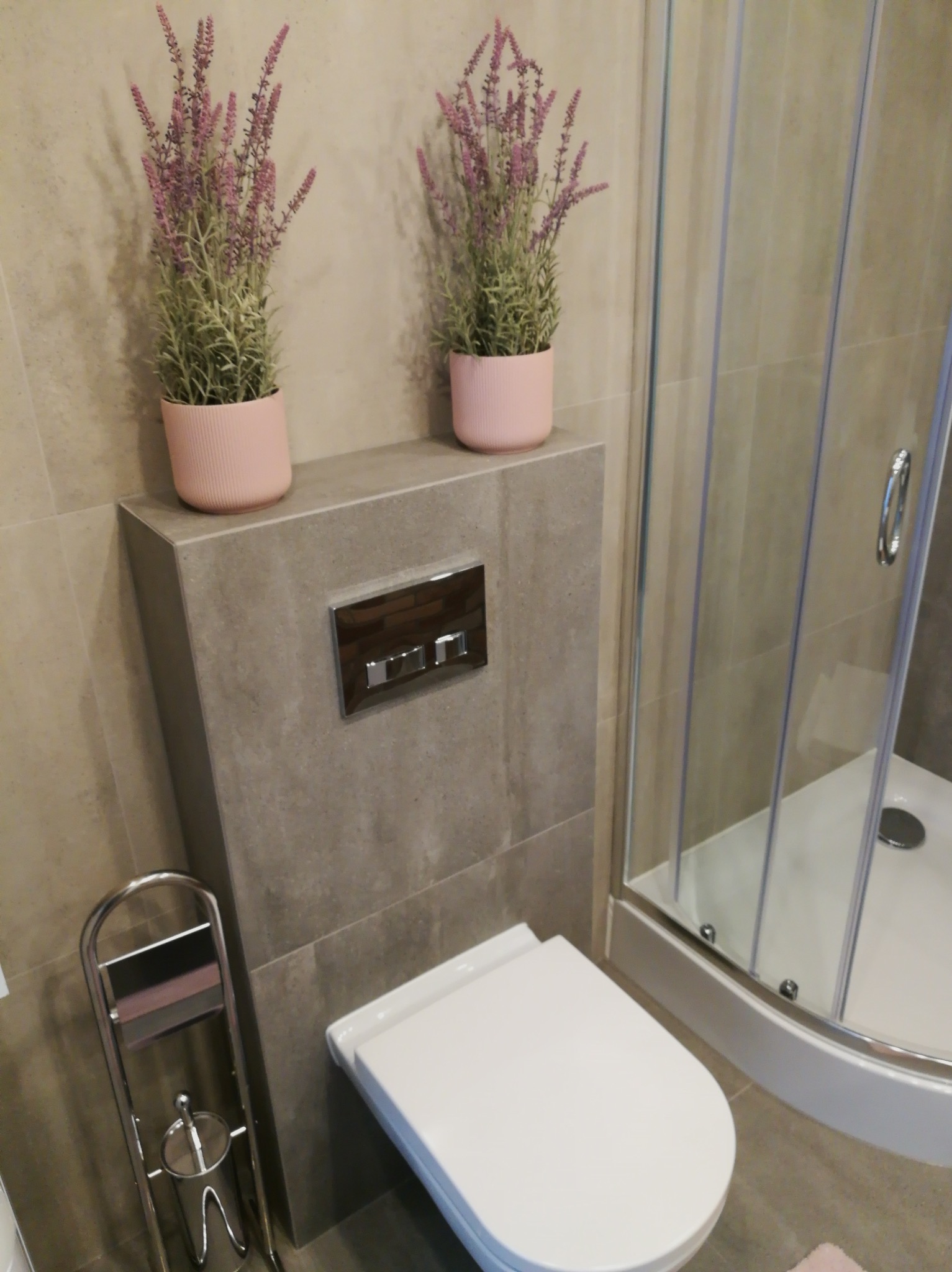 Nowoczesna łazienka z podwieszaną toaletą, szarą obudową i dwoma różowymi doniczkami z lawendą na górze, obok kabiny prysznicowej z przezroczystego szkła.