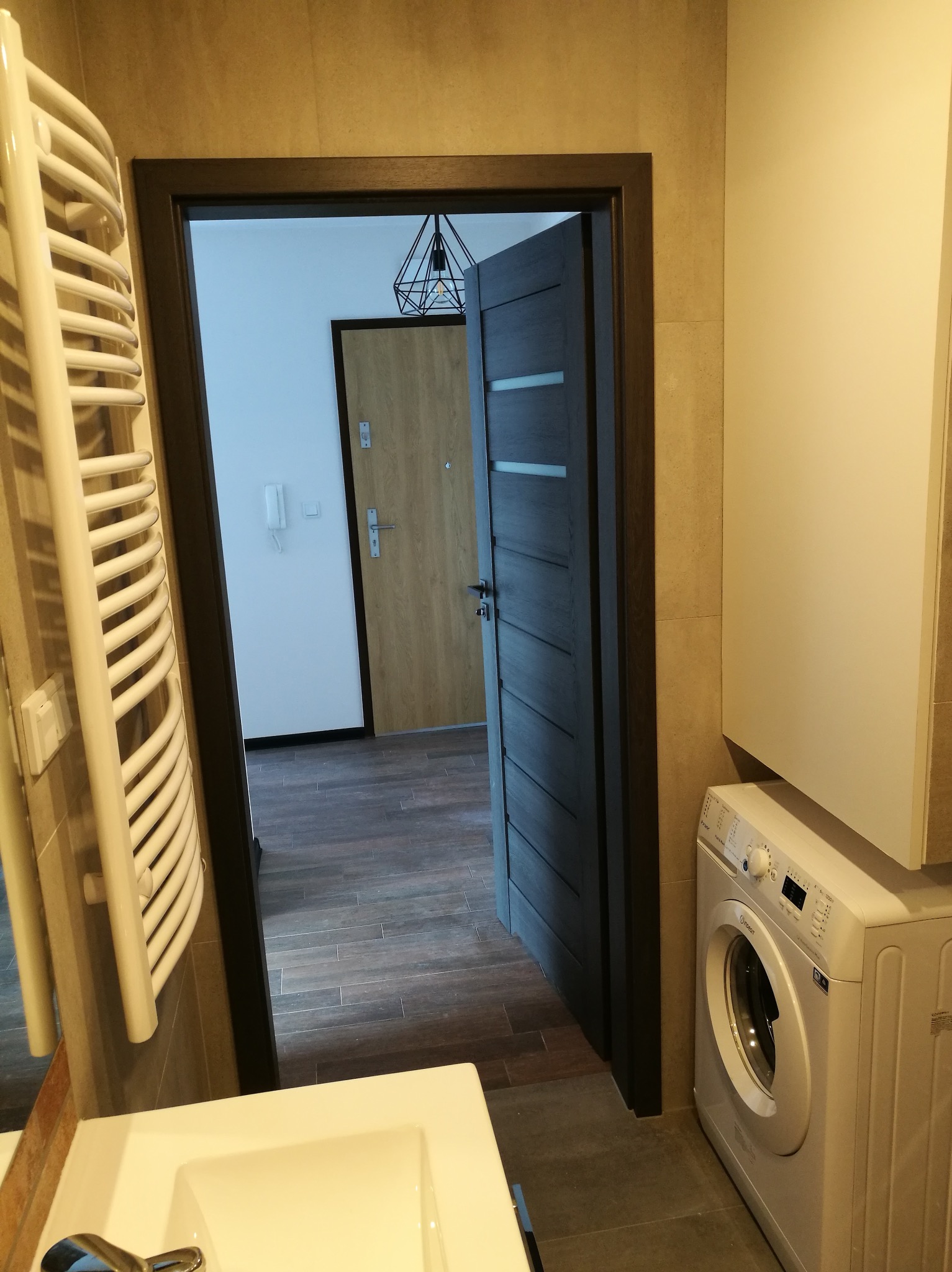 Umeblowana łazienka z pralką, grzejnikiem drabinkowym, umywalką i otwartymi drzwiami do przedpokoju z widoczną lampą wiszącą.