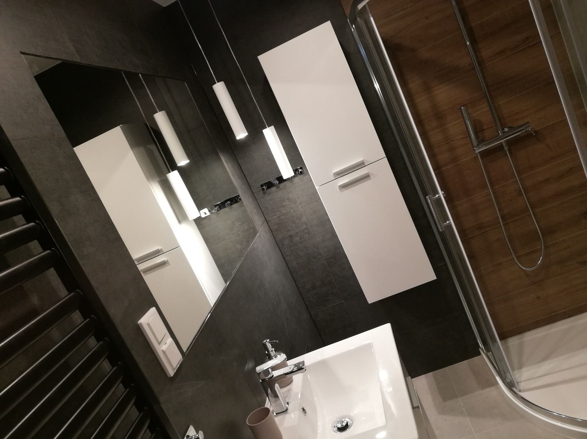 Nowoczesna łazienka z szarą, teksturowaną ścianą, wiszącą białą szafką, podwieszanymi lampami tubami i kabiną prysznicową z drewnopodobną ścianą w tle.