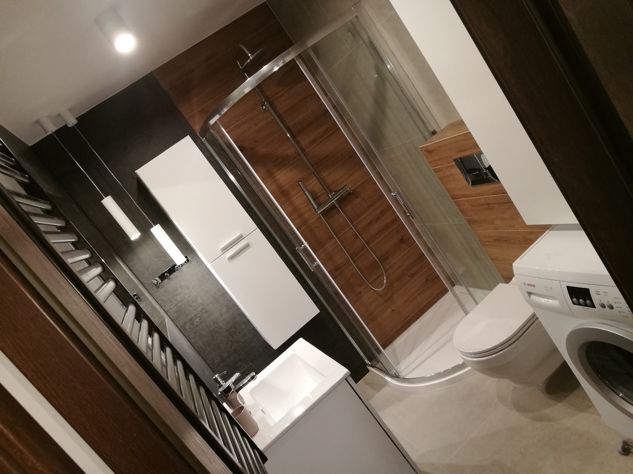 Nowoczesna łazienka z kabiną prysznicową narożną, umywalką nablatową, toaletą wiszącą i pralką Bosch, wykończona płytkami imitującymi drewno i szarym gresem.