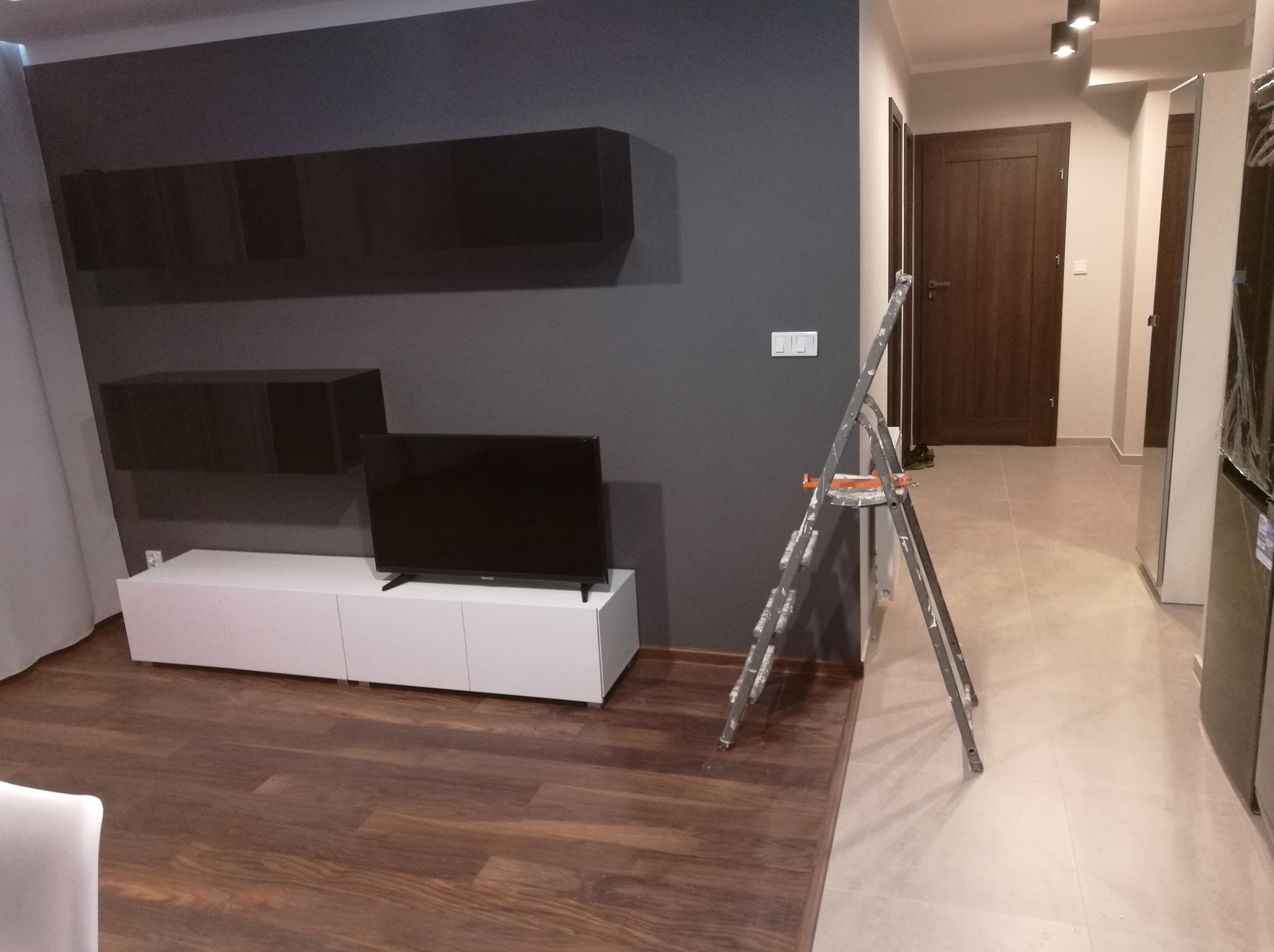 Pokój po świeżym malowaniu ścian na ciemny grafit, widoczna drabina malarska, biała szafka RTV pod telewizorem, drewniana podłoga, drzwi w tle.