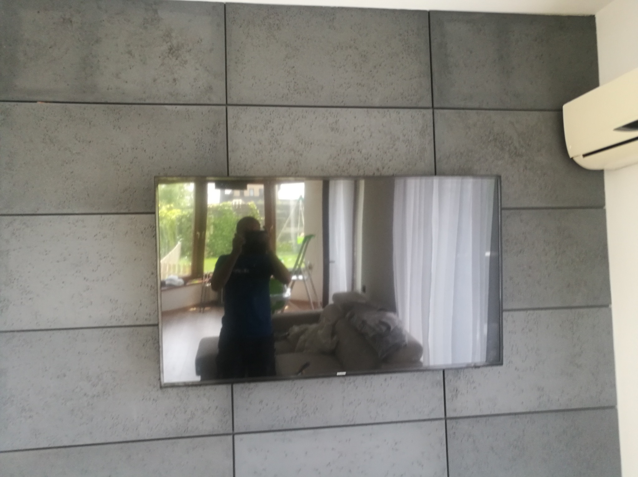 Szara ściana z dekoracyjnych paneli imitujących beton, z zawieszonym telewizorem odbijającym wnętrze pokoju i osobę fotografującą, klimatyzator zamontowany na górze po prawej stronie.