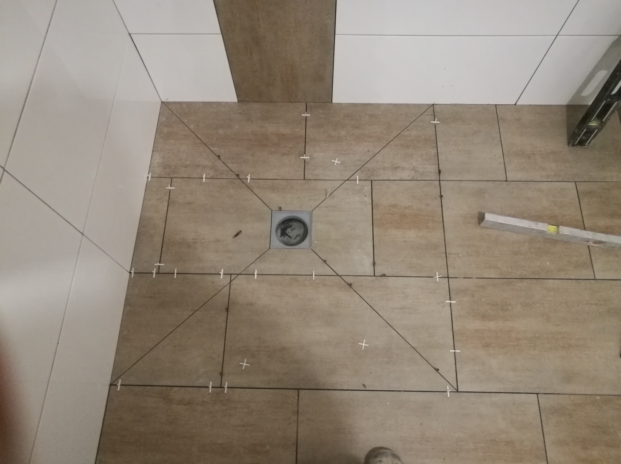 Układanie brązowych płytek podłogowych w łazience z widocznym odpływem liniowym i krzyżykami dystansowymi, częściowo wykończone białe płytki ścienne.