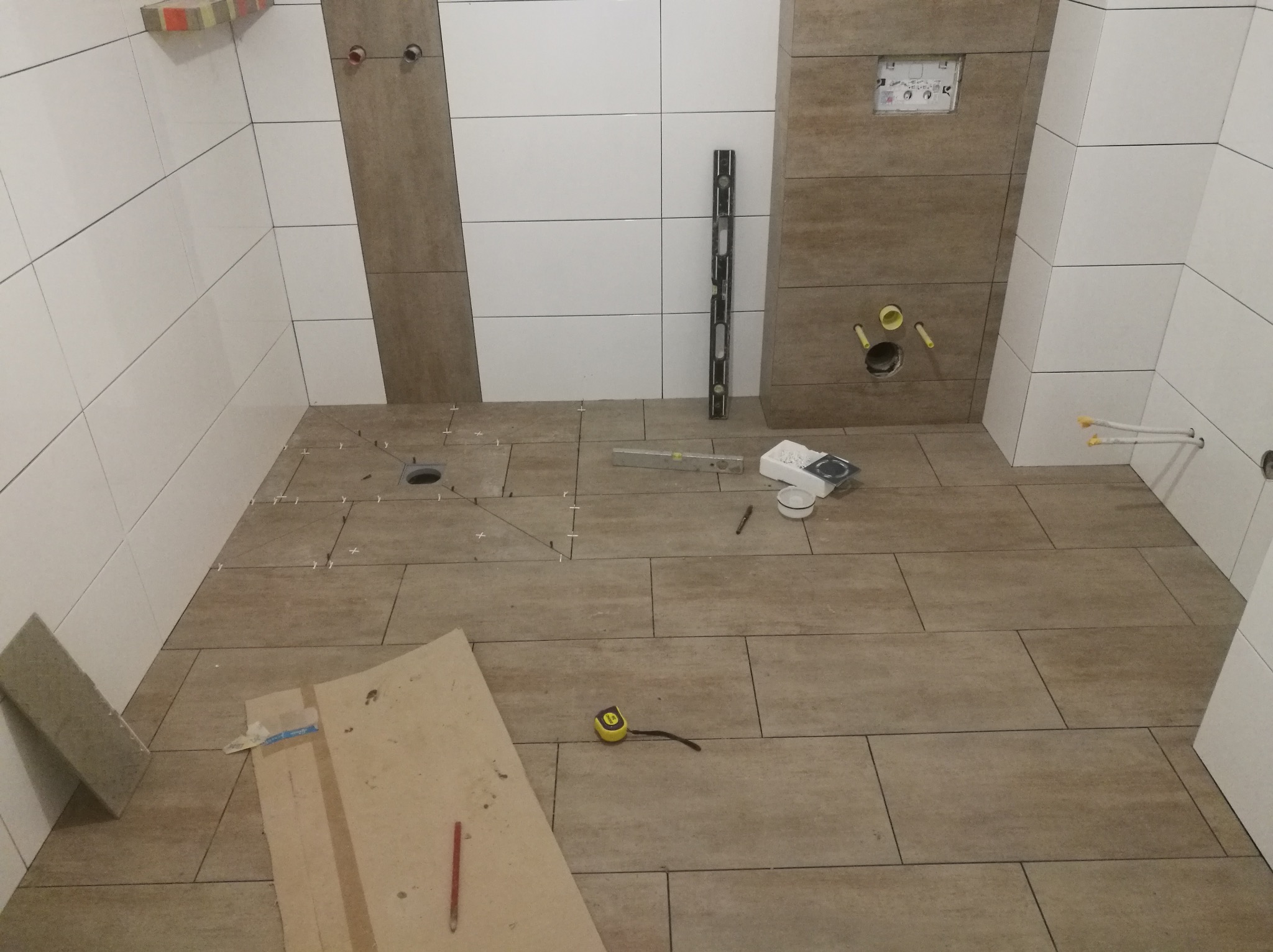 Wnętrze łazienki w trakcie układania podłogowych płytek ceramicznych imitujących drewno, widoczne narzędzia: poziomica, miara zwijana, ołówek, krzyżyki dystansowe, płyta kartonowa z naniesionymi...