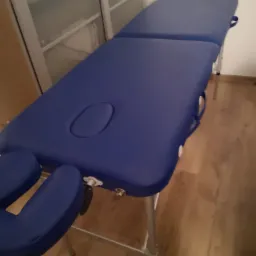 Niebieski stół do zabiegów z regulowanym zagłówkiem, widoczny w neutralnym pomieszczeniu z drewnianą podłogą i jasną ścianą.