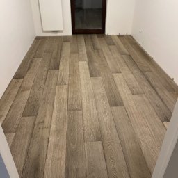 Project MasterWood Przemysław Bejmert - Ułożona podłoga z paneli imitujących drewno w pustym, jasnym pokoju, z widocznymi dystansami przy ścianach i ciemnym oknem w tle.