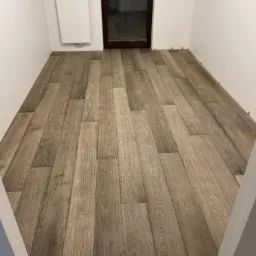 Ułożona podłoga z paneli imitujących drewno w pustym, jasnym pokoju, z widocznymi dystansami przy ścianach i ciemnym oknem w tle.