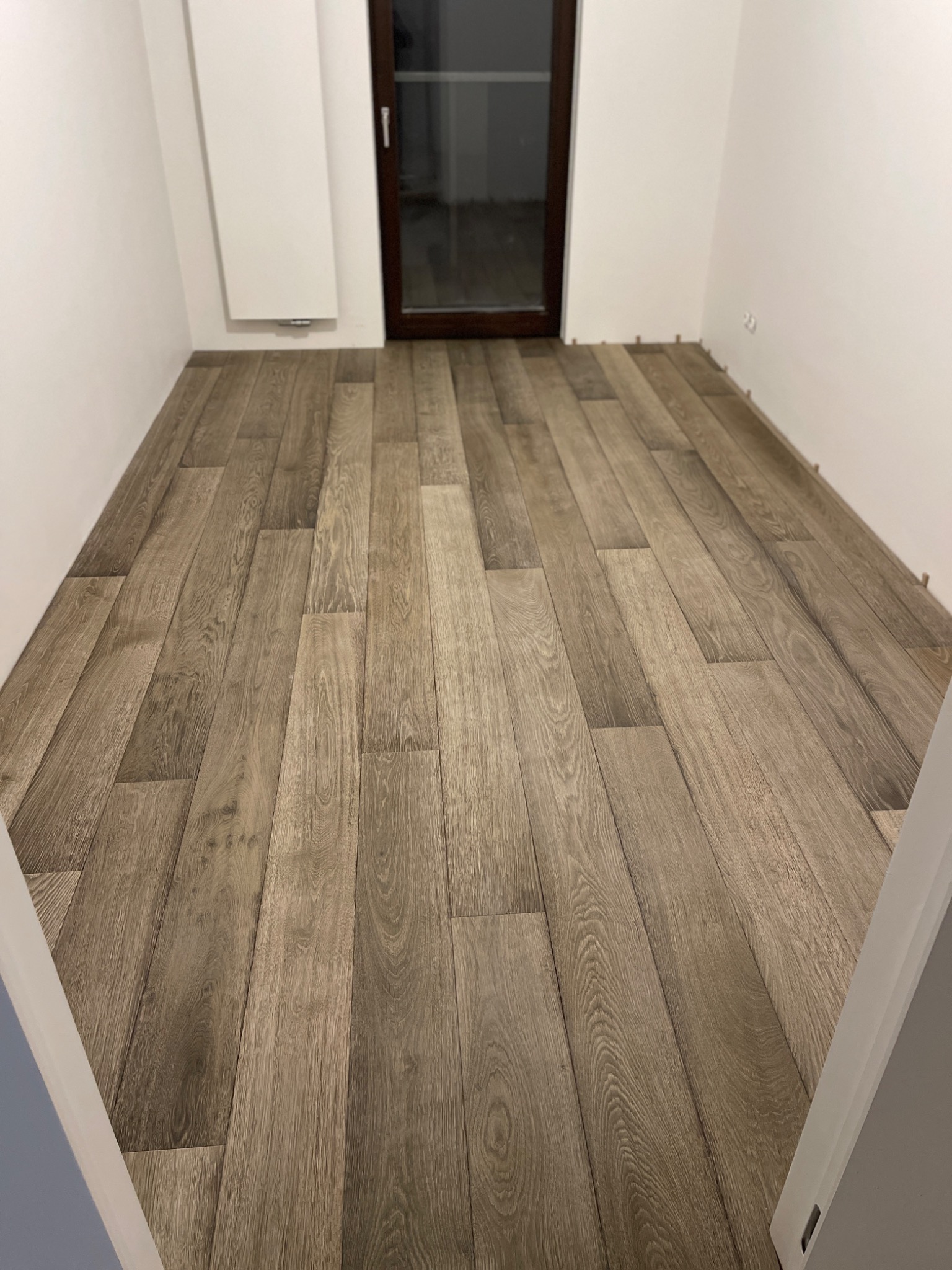 Ułożona podłoga z paneli imitujących drewno w pustym, jasnym pokoju, z widocznymi dystansami przy ścianach i ciemnym oknem w tle.