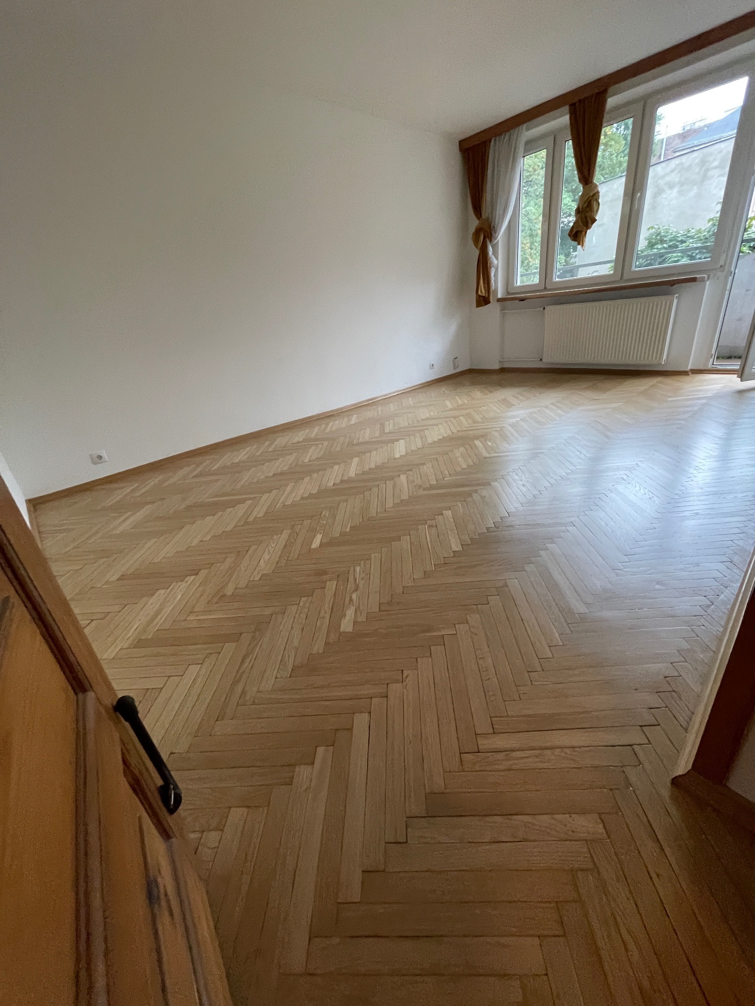 Wycyklinowany parkiet ułożony w jodełkę francuską, widok z perspektywy wejścia do pokoju, naturalne oświetlenie z okna z dekoracyjnymi zasłonami.