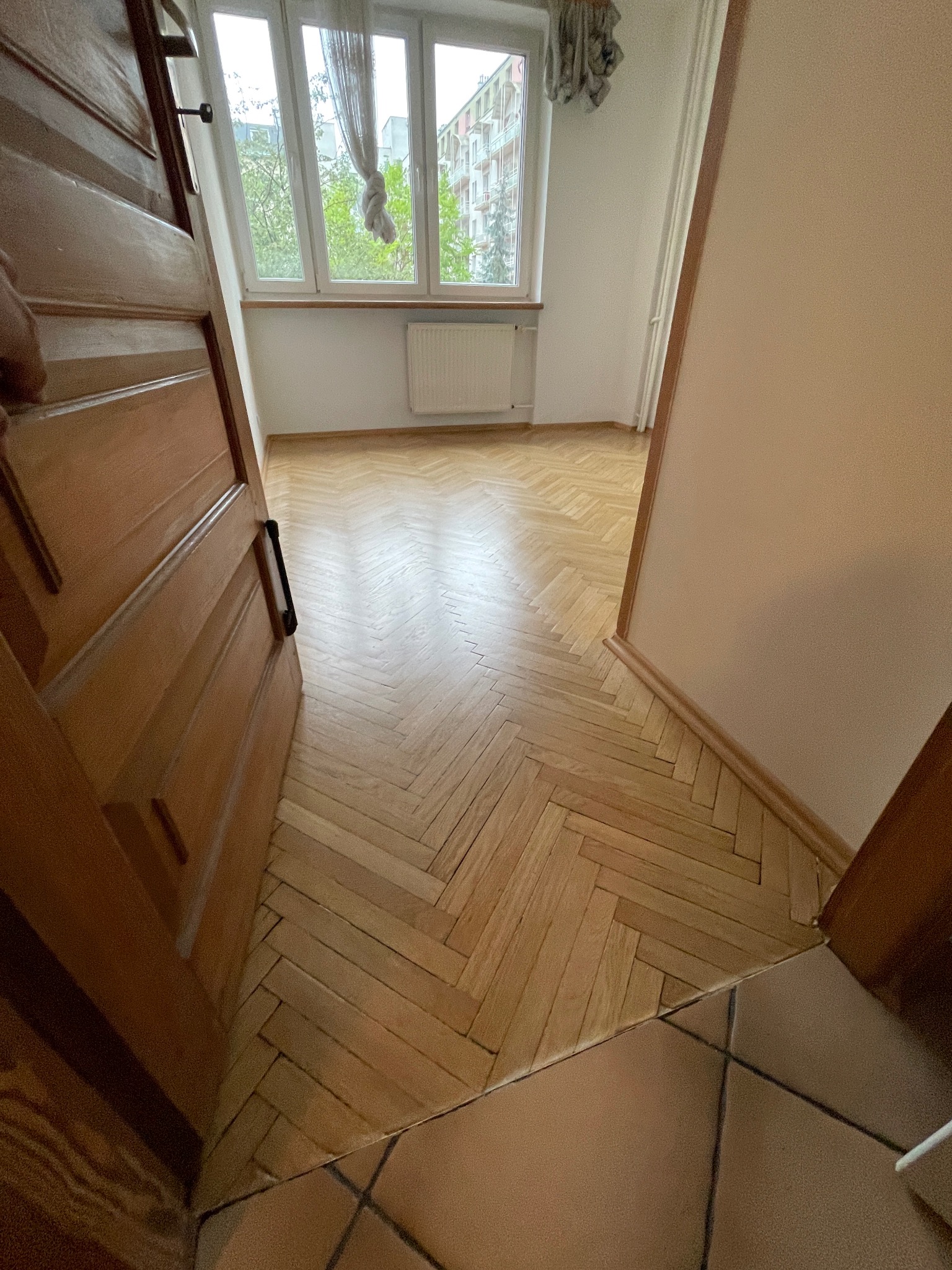 Widok zza uchylonych drzwi na jasny parkiet ułożony w jodełkę w pokoju z oknem i grzejnikiem. Część podłogi wyłożona brązowymi płytkami.