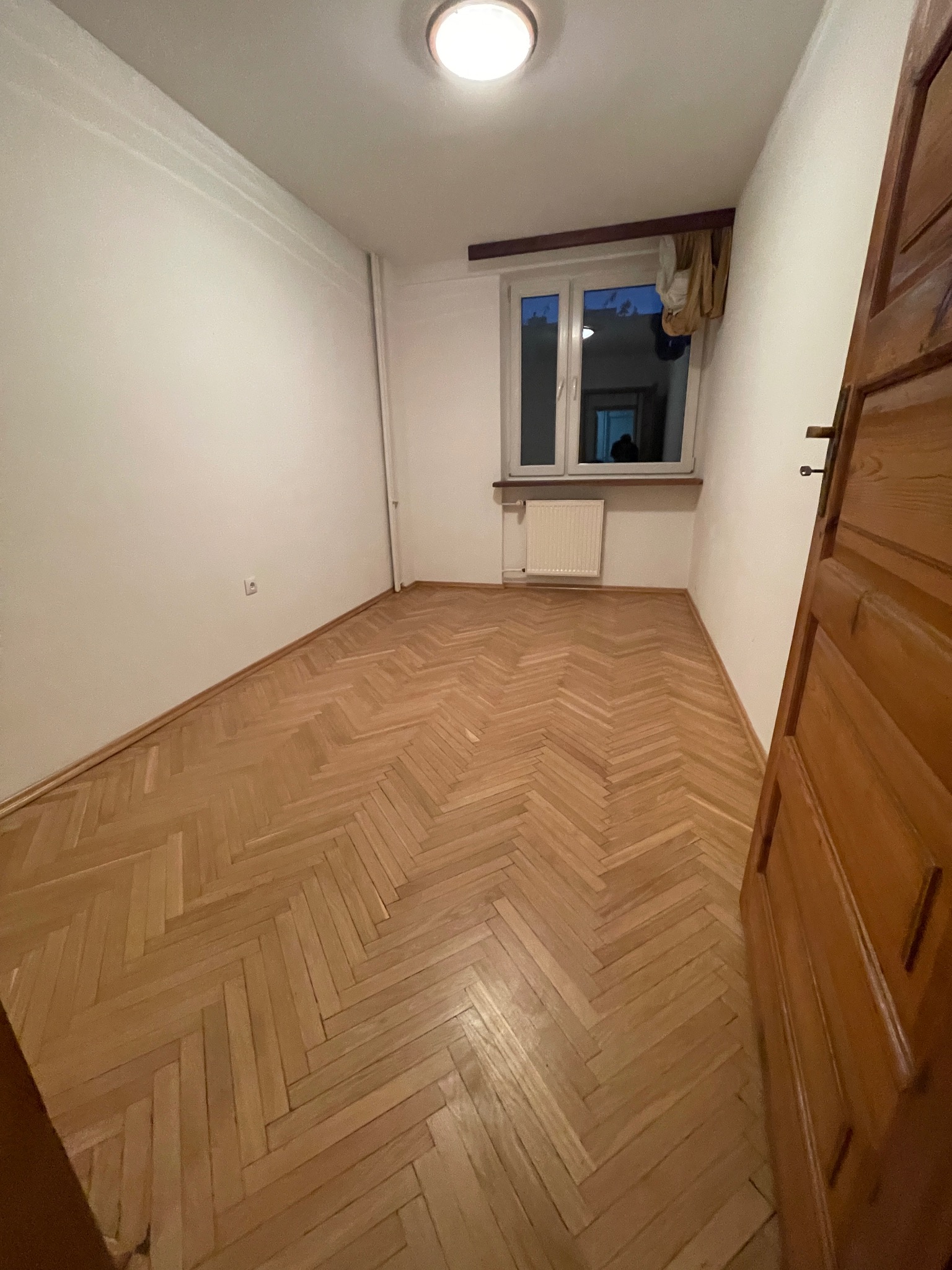 Pokój z parkietem ułożonym w jodełkę, widoczne białe ściany, okno z grzejnikiem i fragment drewnianych drzwi po prawej stronie kadru.
