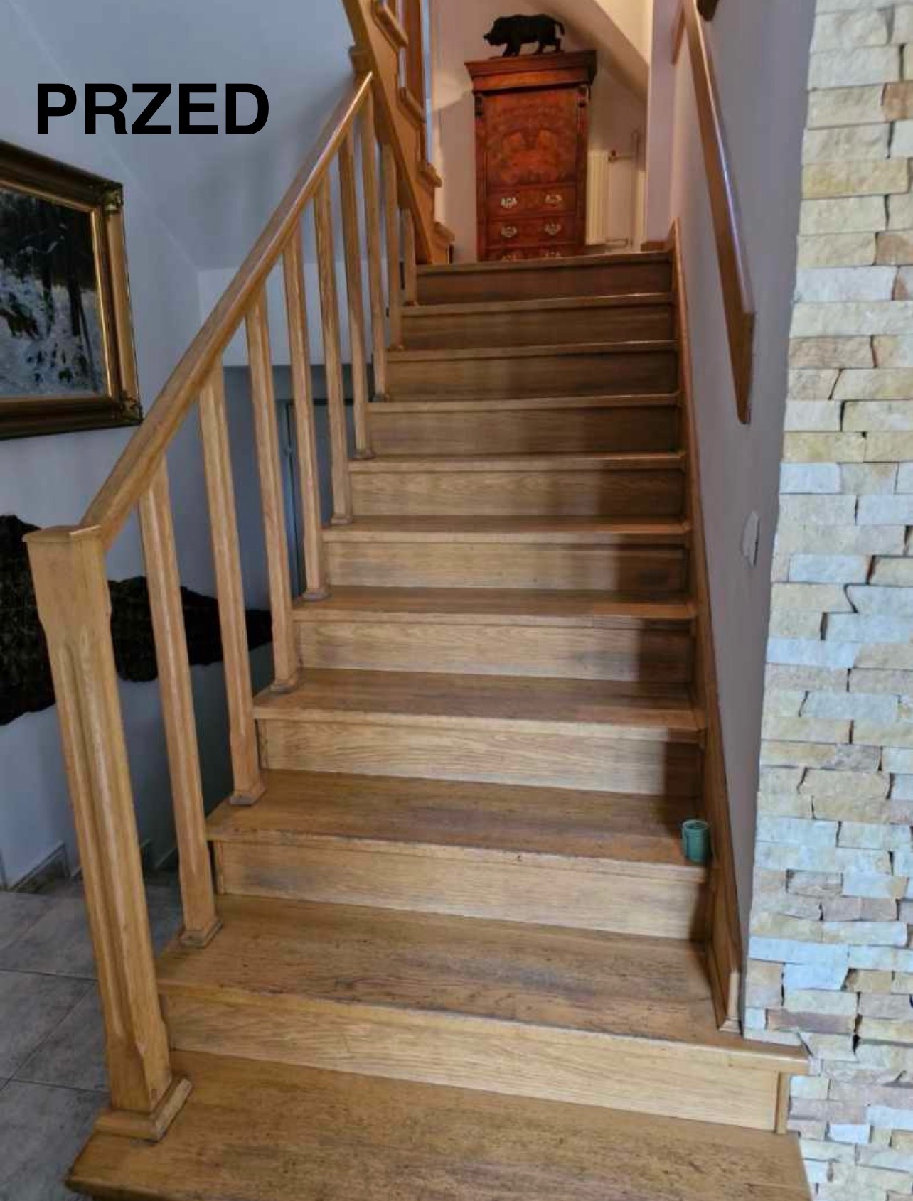 Drewniane schody z jasnymi balustradami przed cyklinowaniem, widoczne ślady użytkowania. Na ścianie dekoracyjna okładzina z kamienia. U góry szafa z rzeźbą dzika.