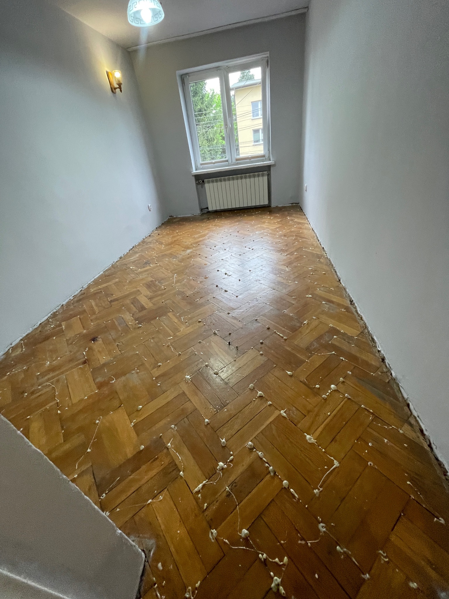 Parkiet ułożony w jodełkę przed renowacją, z widocznymi śladami po kleju i szpachli. W tle okno z widokiem na budynek i grzejnik.