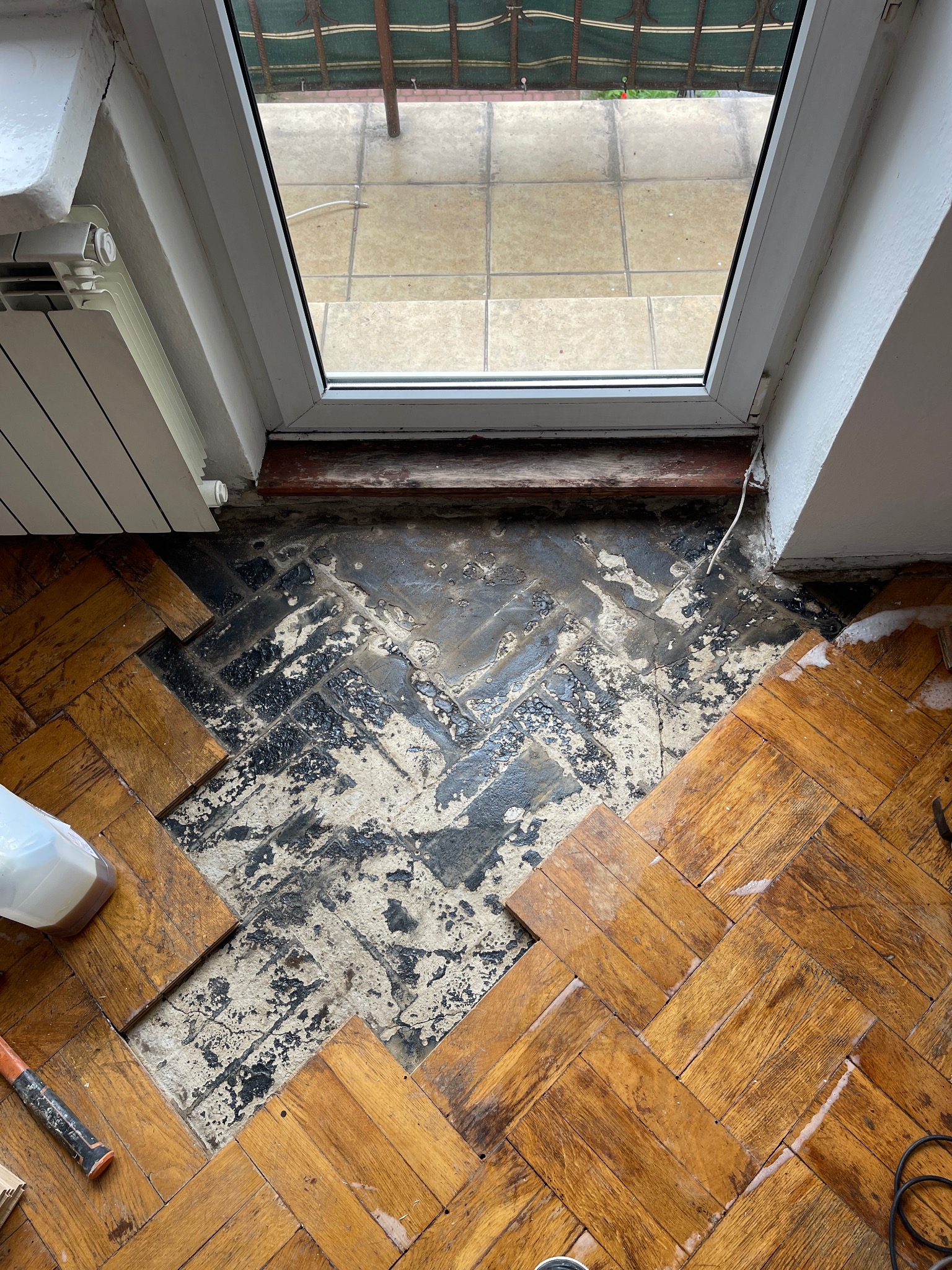 Fragment podłogi z ułożonym parkietem w jodełkę, częściowo uszkodzonym. Widoczne narzędzia i chemia. W tle okno balkonowe i grzejnik.