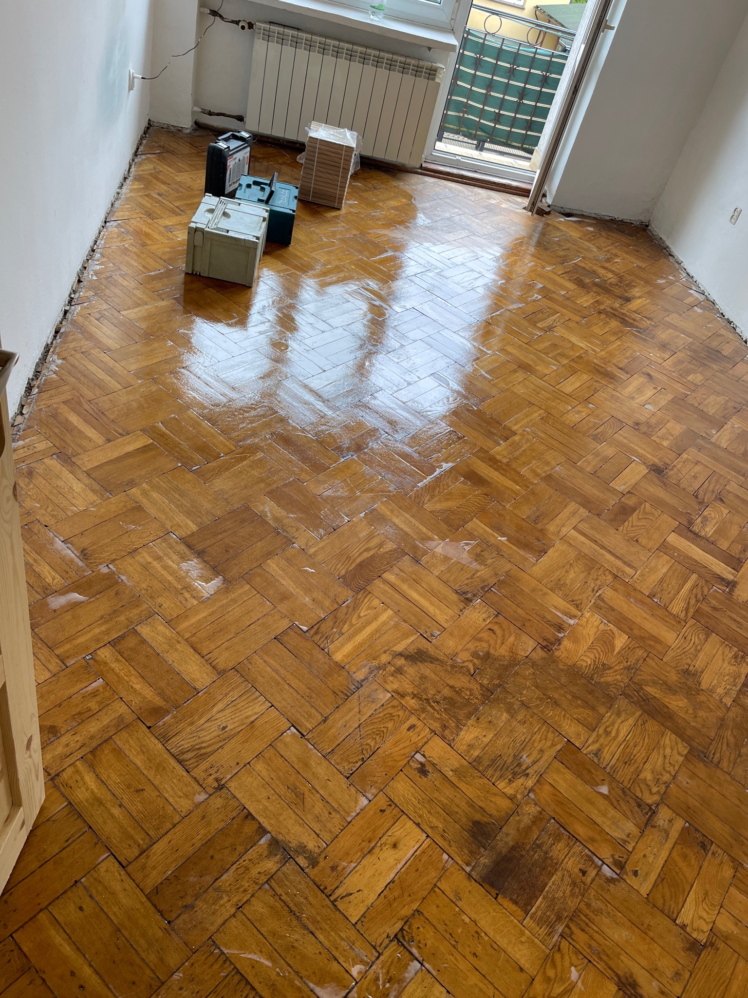 Parkiet w jodełkę, częściowo wyczyszczony, z widocznymi narzędziami i otwartym balkonem w tle. Prace renowacyjne w toku, widoczne ślady po czyszczeniu.