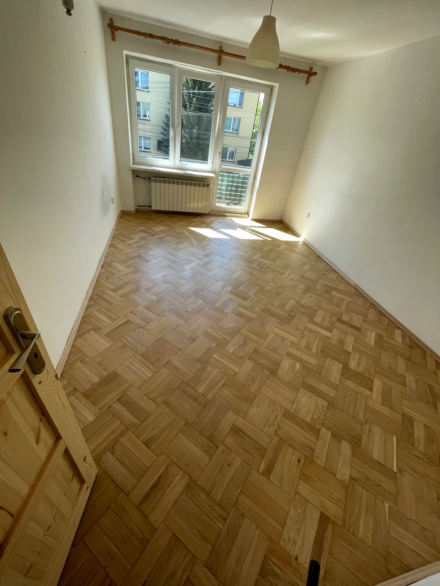 Jasny parkiet ułożony w jodełkę w pustym pokoju z oknem balkonowym i grzejnikiem. Widoczny fragment drewnianych drzwi po lewej stronie kadru.