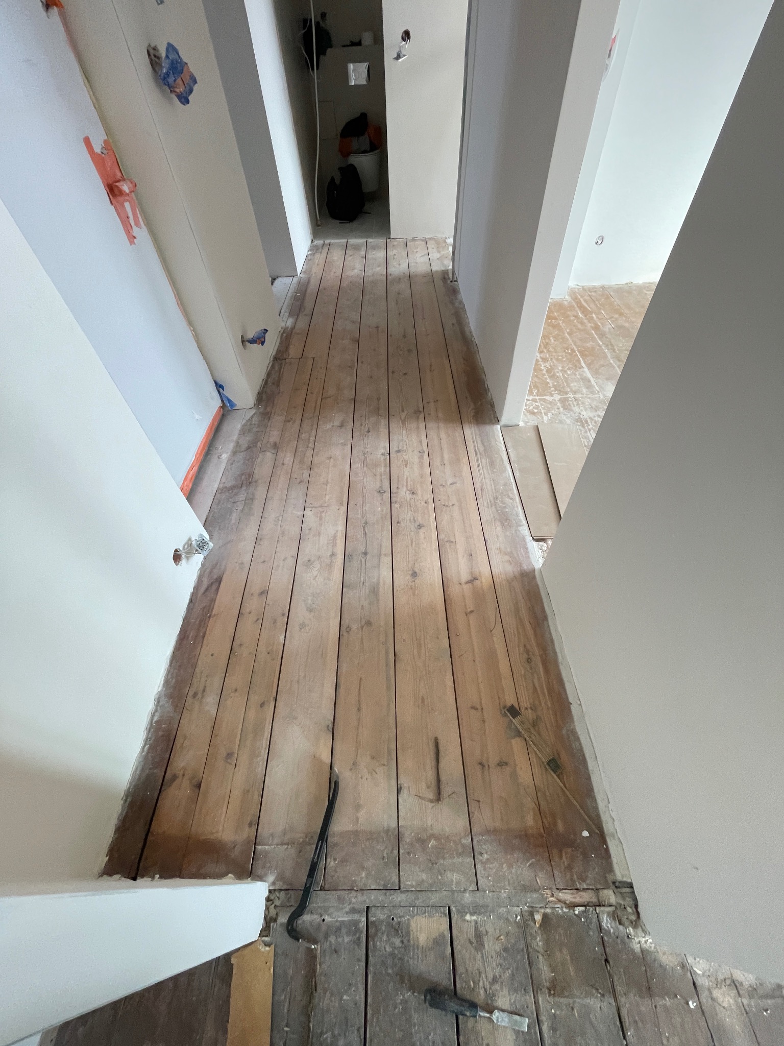 Korytarz z drewnianą podłogą w trakcie renowacji. Widoczne narzędzia i fragmenty starej podłogi. Świeżo pomalowane ściany i zabezpieczenia.