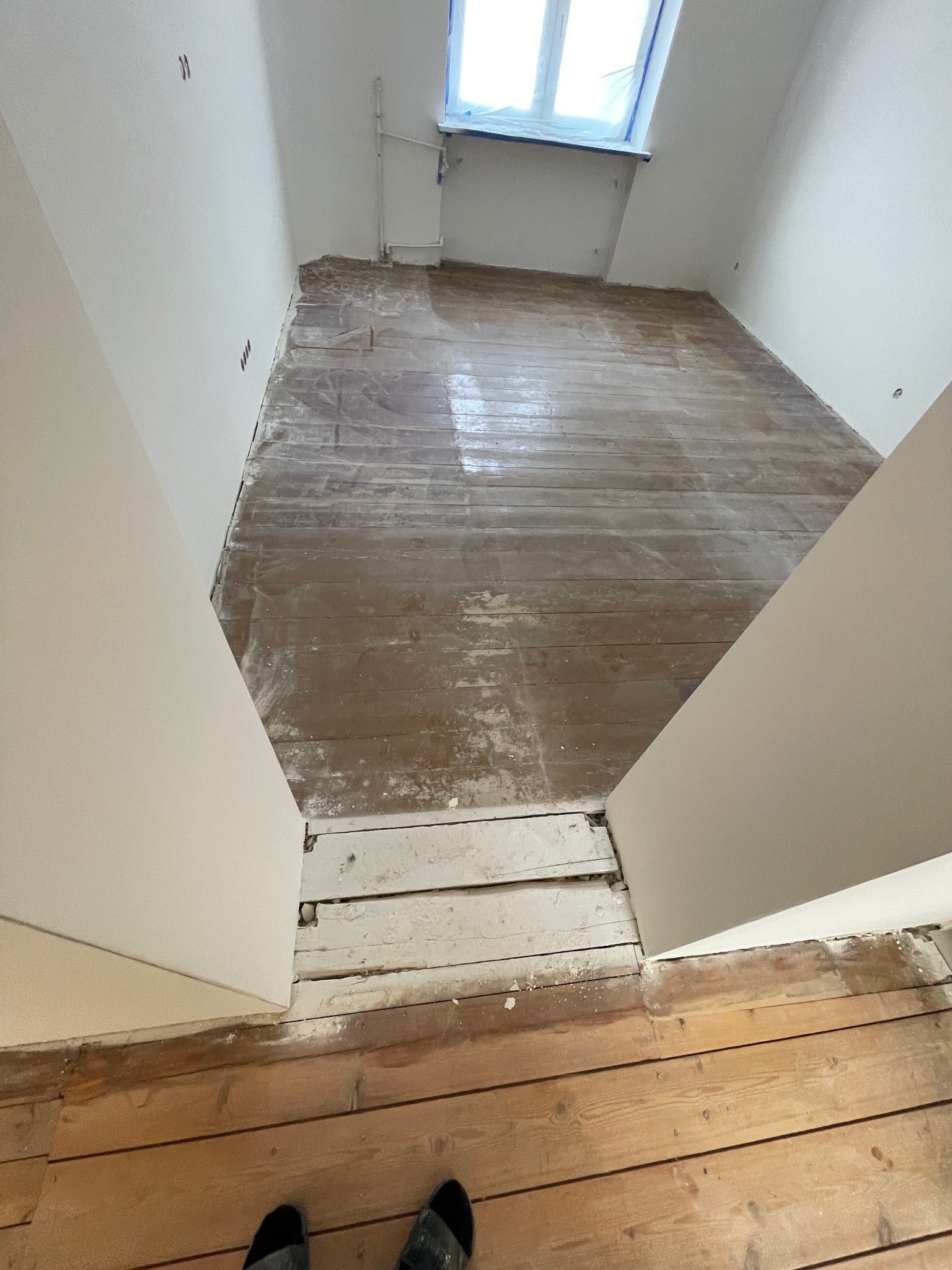 Pokój z podłogą w trakcie renowacji. Część desek po obróbce, część jeszcze nie. Widoczne ślady pyłu i niedoskonałości. Okno zabezpieczone folią.