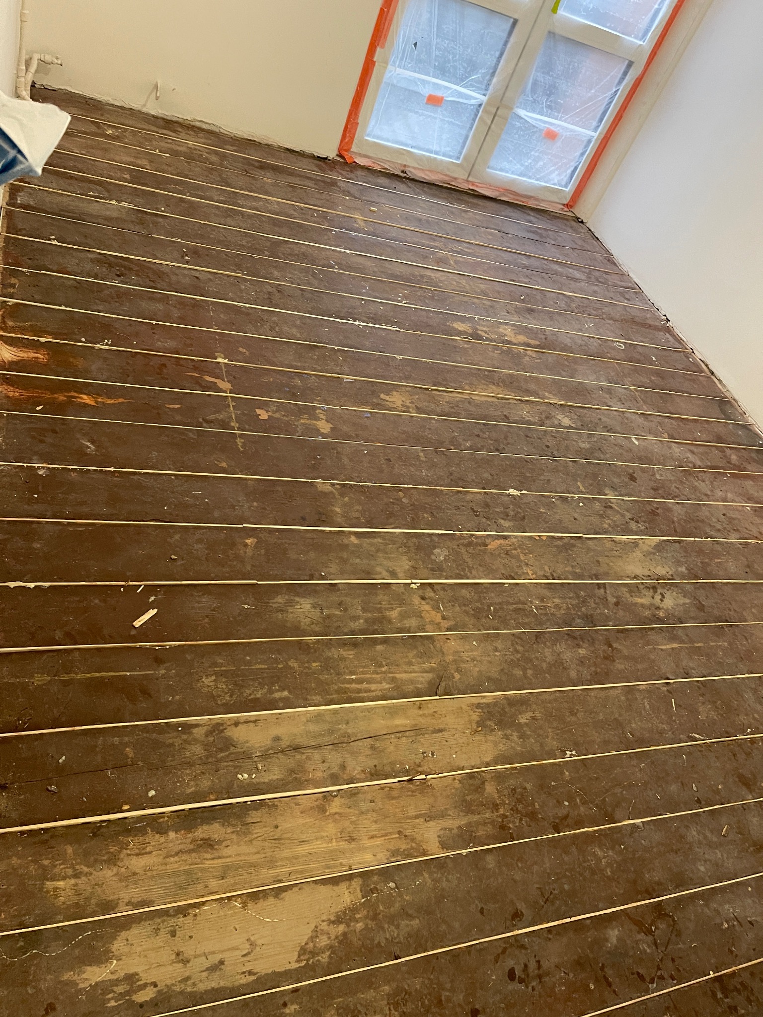 Parkiet przed renowacją: widoczne stare deski z ubytkami i resztkami farby, okno zabezpieczone folią. Perspektywa od podłogi w górę, surowe oświetlenie.