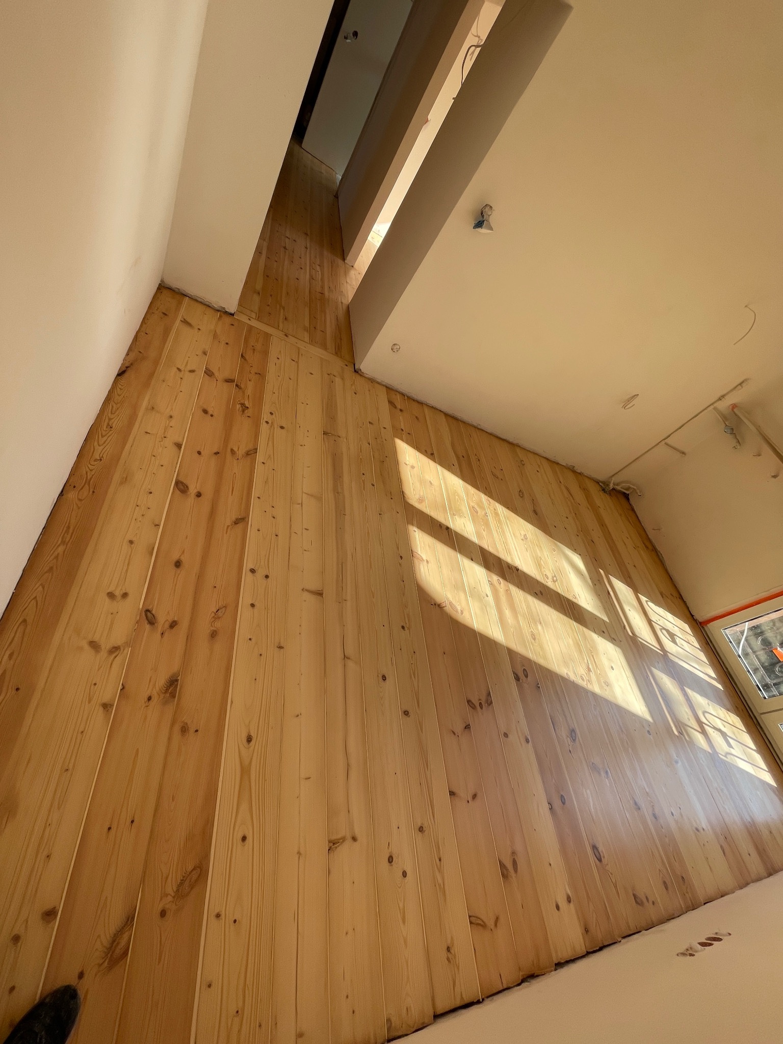 Jasne, drewniane deski podłogowe w nowym pomieszczeniu, oświetlone naturalnym światłem słonecznym wpadającym przez okno. Widoczny fragment buta w rogu kadru.