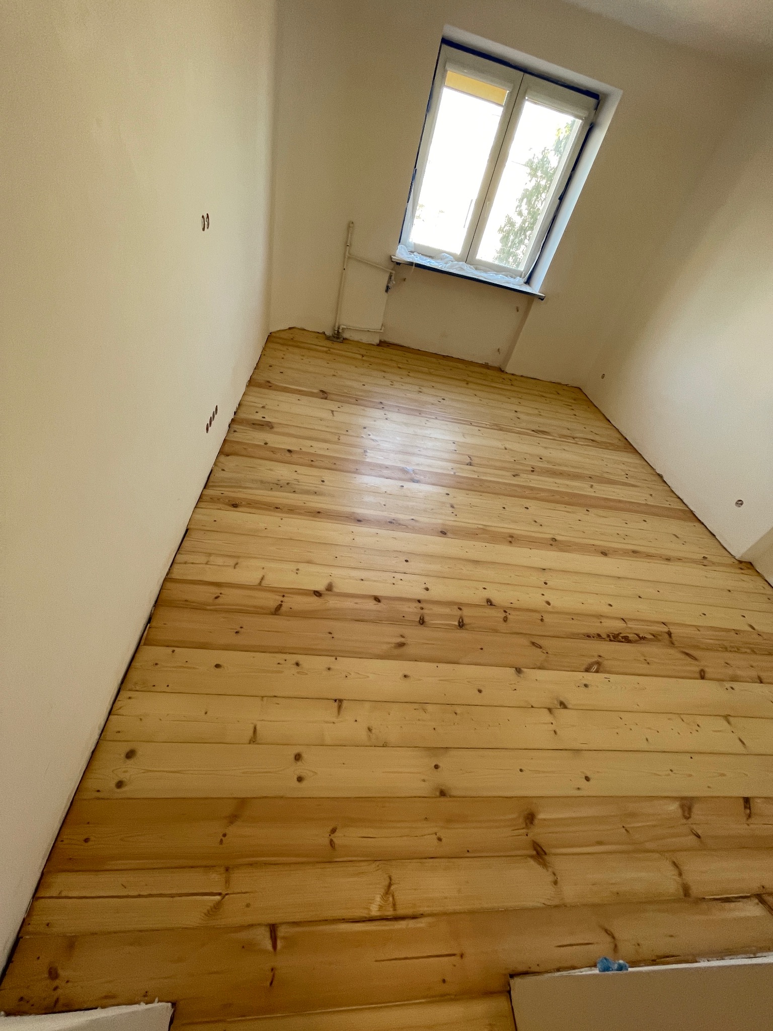 Wykończona podłoga z jasnego drewna w pokoju z oknem dachowym. Widoczne deski podłogowe po renowacji, z naturalnym rysunkiem drewna i połyskiem lakieru.