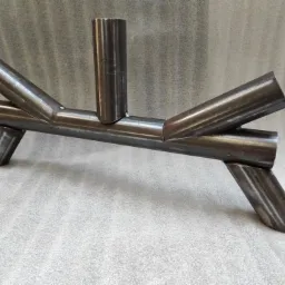 Surowa, spawana konstrukcja z rur stalowych o ciemnym, metalicznym połysku, ustawiona na szarym tle, prezentująca połączenia spawane i surowe krawędzie cięcia.