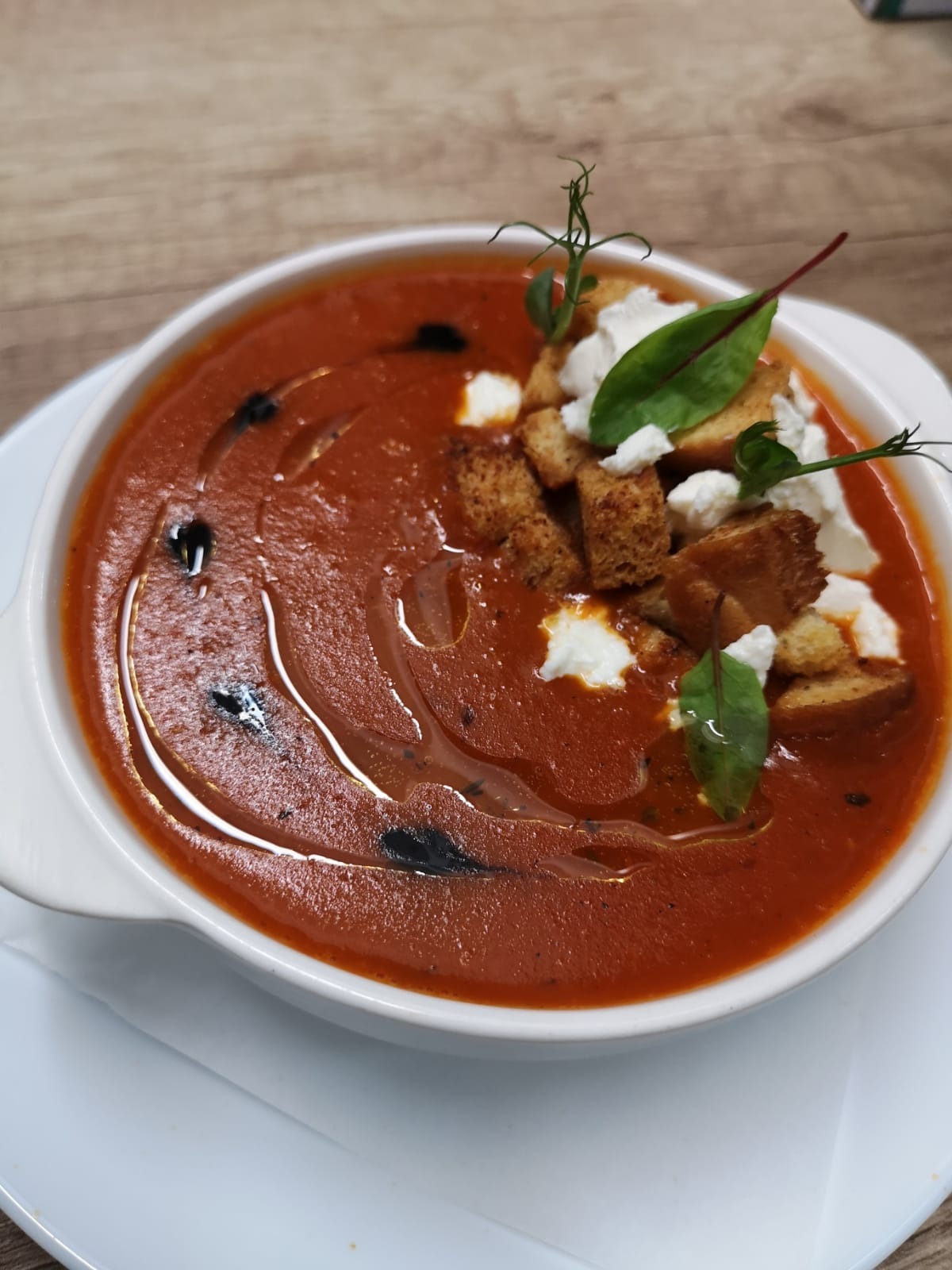 Krem pomidorowy w białej miseczce, udekorowany grzankami, kremowym serem i świeżymi listkami z kroplami oliwy.