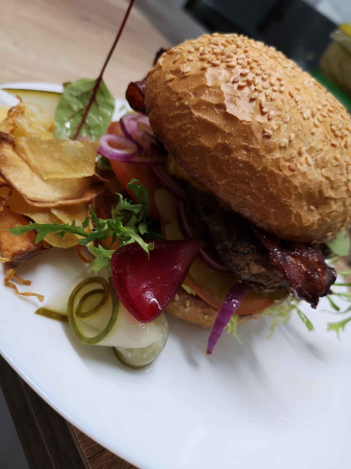 Apetyczny burger z wołowiną, boczkiem, warzywami i sosem, podany z chipsami i ogórkami konserwowymi na białym talerzu.