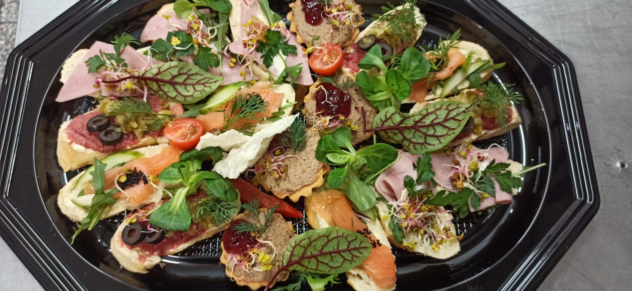 Czarny, plastikowy talerz z różnorodnymi kanapkami i tartaletkami, udekorowanymi świeżymi ziołami, warzywami i kiełkami.