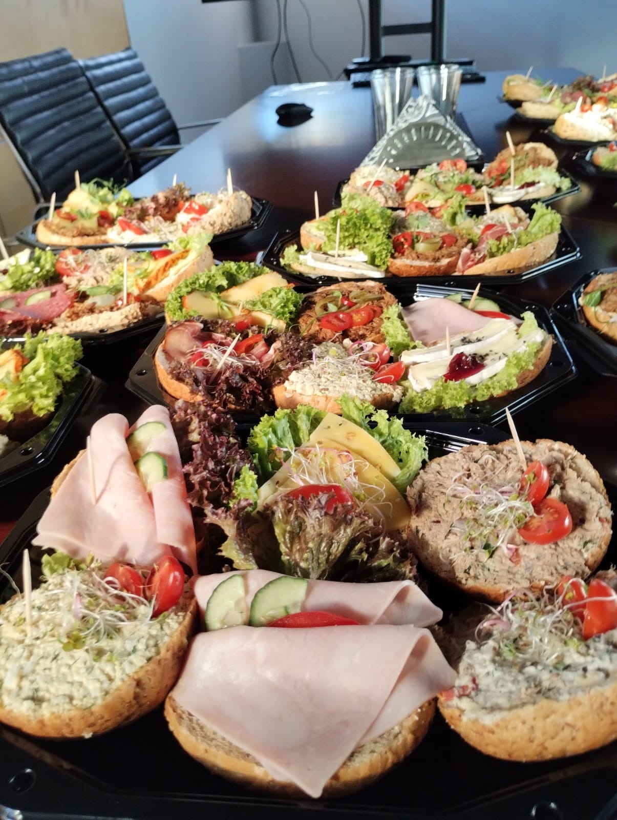 Różnorodne kanapki na pieczywie, udekorowane warzywami i wędlinami, podane na czarnych tackach na ciemnym stole w biurowym otoczeniu.