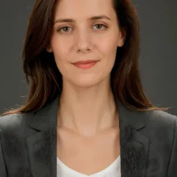 Portret uśmiechniętej kobiety w szarym żakiecie i białej bluzce na jednolitym, ciemnoszarym tle.