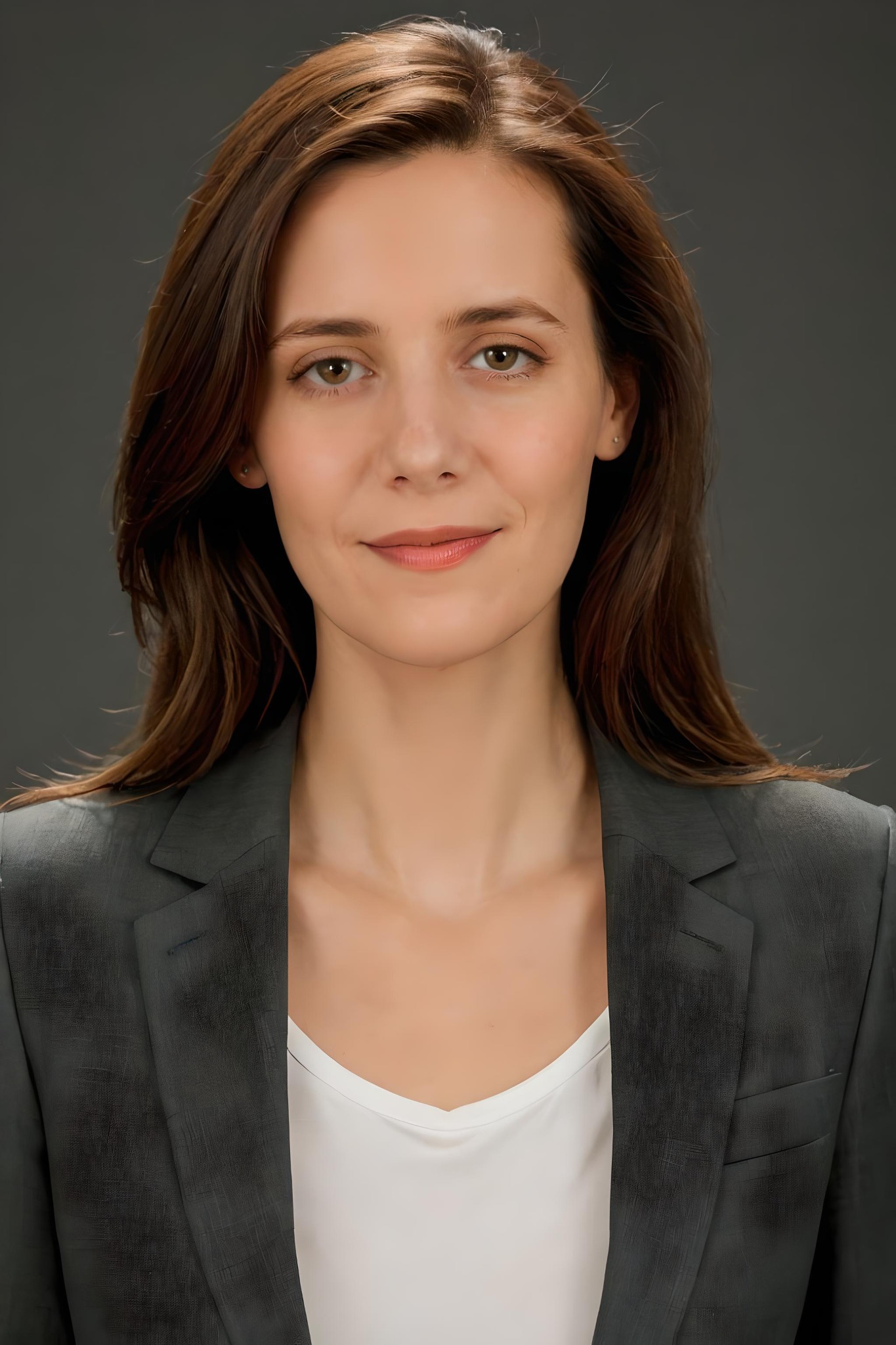 Portret uśmiechniętej kobiety w szarym żakiecie i białej bluzce na jednolitym, ciemnoszarym tle.