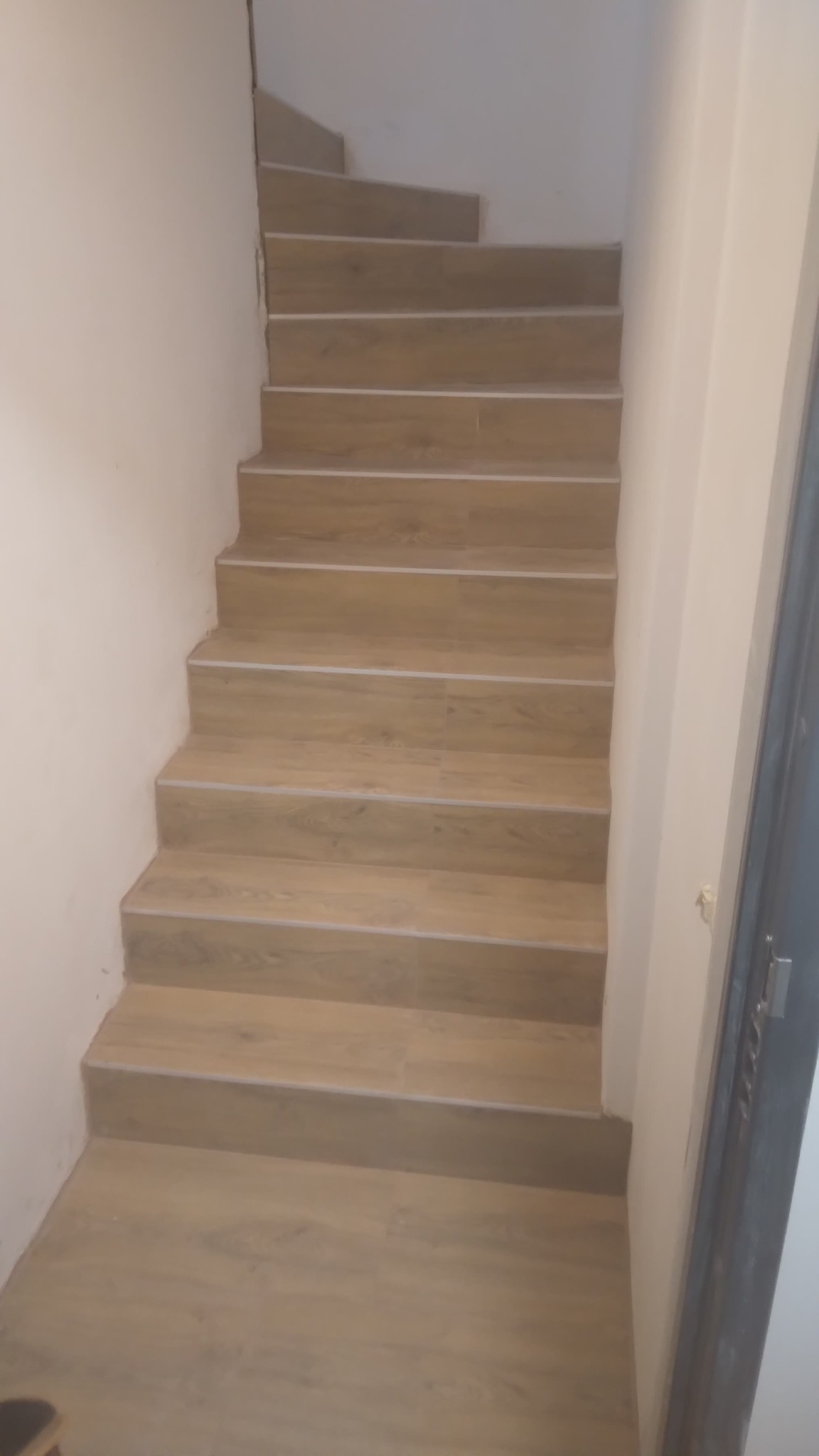 Schody wyłożone płytkami imitującymi drewno, z delikatnymi białymi fugami, prowadzące w górę w jasnym, minimalistycznym wnętrzu.