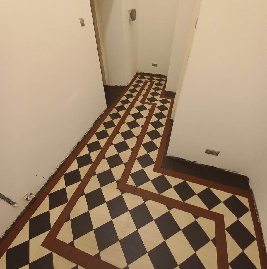 Ułożona podłoga z płytek w szachownicę (czarne i kremowe) z brązowymi obramowaniami w korytarzu, widok z góry, ściany białe.