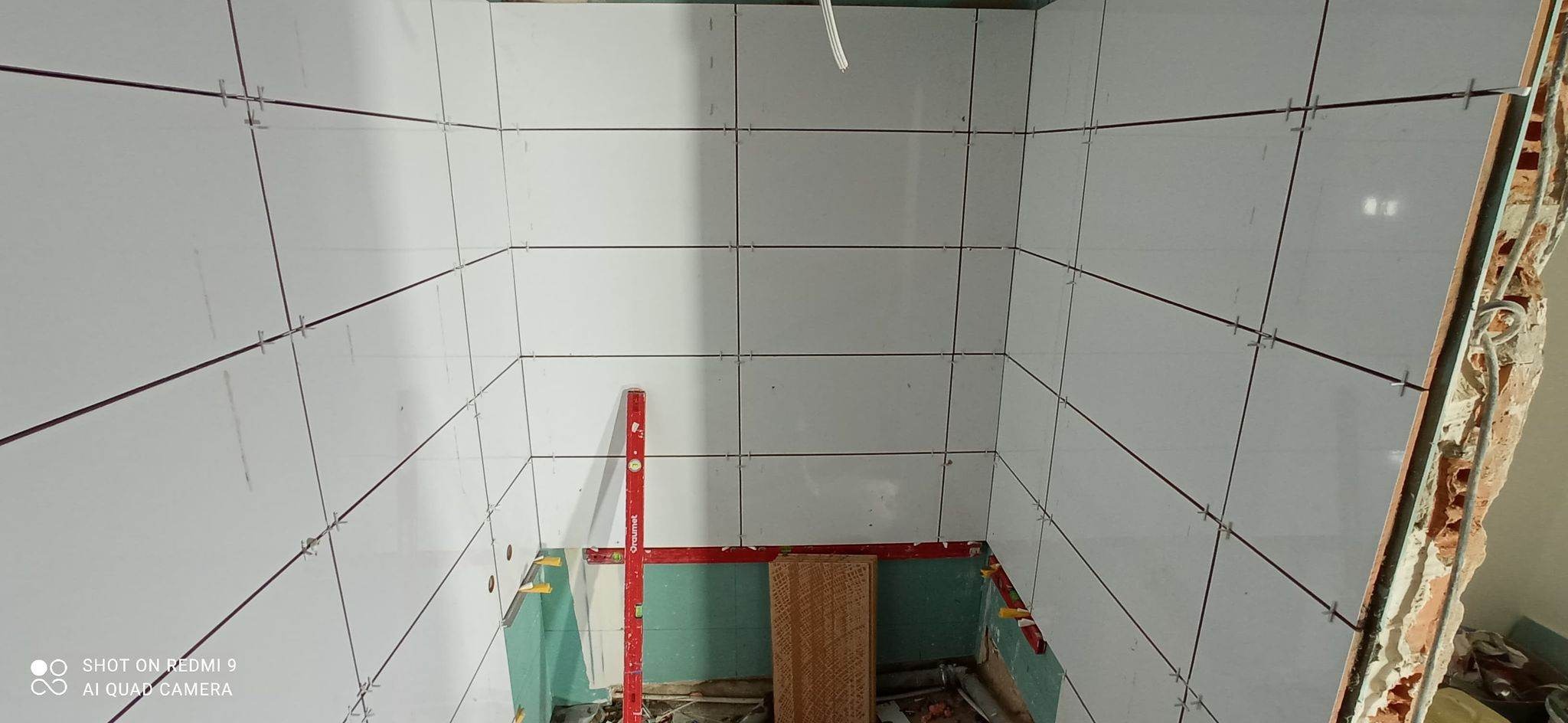 Wnętrze łazienki w trakcie układania białych płytek, widoczne krzyżyki dystansowe, poziomica i fragment odsłoniętej ściany z cegły.