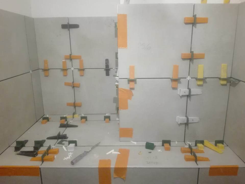 Układanie szarych płytek z użyciem pomarańczowych i żółtych klinów poziomujących oraz czarnych krzyżyków dystansowych, widoczny nóż do cięcia płytek.