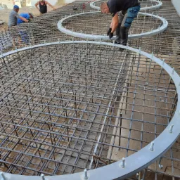 Prace zbrojarskie: Widok z góry na siatkę z prętów zbrojeniowych, z trzema okrągłymi elementami mocującymi, na placu budowy z pracownikami w tle oraz stertą ziemi.