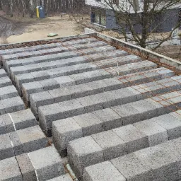 Wznoszenie ścian z szarej bloczków betonowych na ruszcie z prętów zbrojeniowych, widoczne spoiny wypełnione pianką montażową, w tle budynek w stanie surowym otoczony drzewami.