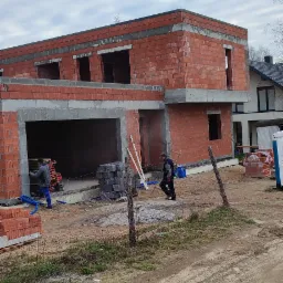 Wznoszony dwukondygnacyjny budynek z czerwonej cegły, z widocznymi otworami okiennymi i garażem w stanie surowym, dwóch robotników przy pracy, stosy bloczków i cegieł na placu budowy.