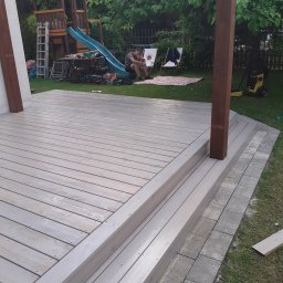 Sauny - Świeżo pomalowany drewniany taras z dwoma stopniami, widoczne słupy konstrukcyjne, w tle plac zabaw dla dzieci i osoba siedząca na trawie.